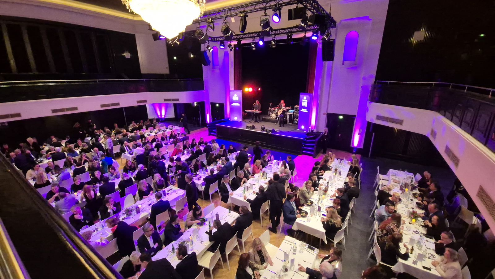 Festlicher Ballsaal mit langen Tafeln, Bühne und violetter Lichtstimmung
